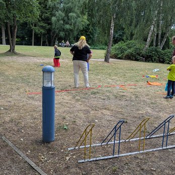 zeigt Kind beim Sackhüpfen auf einer Wiese und eine beobachtende Frau