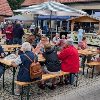 zeigt Markttreiben mit stehenden und an Holzgarnituren sitzenden Menschengruppen