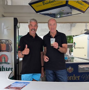 zeigt 2 Männer an einem Bierstand mit Becher mit Logo der Blaubeerstadt