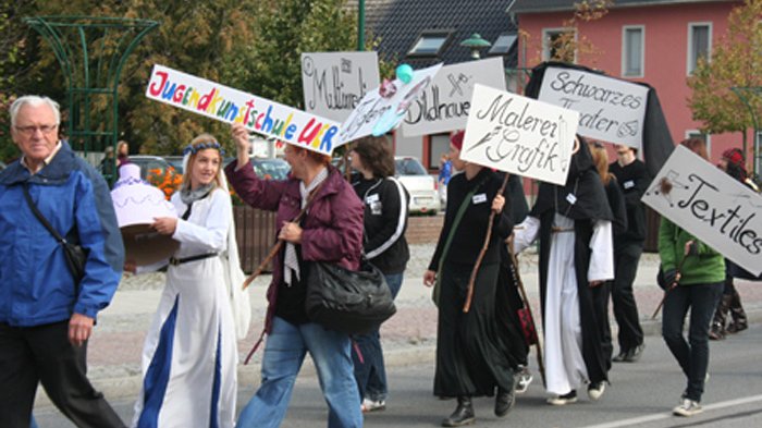 zeigt Ausschnitt des Festumzugs mit kostümierten Personen der Jugendkunstschule UER, die Schilder hochhalten