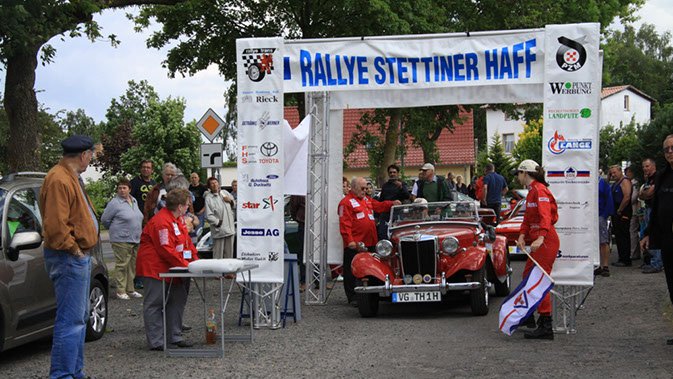 zeigt Starttor der Rallye mit einem abfahrbereiten Oldtimer und Rallyepersonal