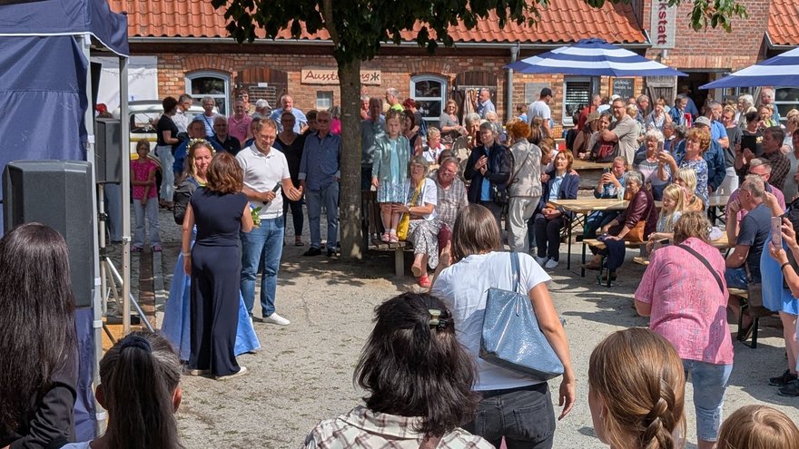 zeigt Blick auf den Festplatz mit einer großen Menschengruppe, die Personen vor einer Bühne zuhören und zuschauen